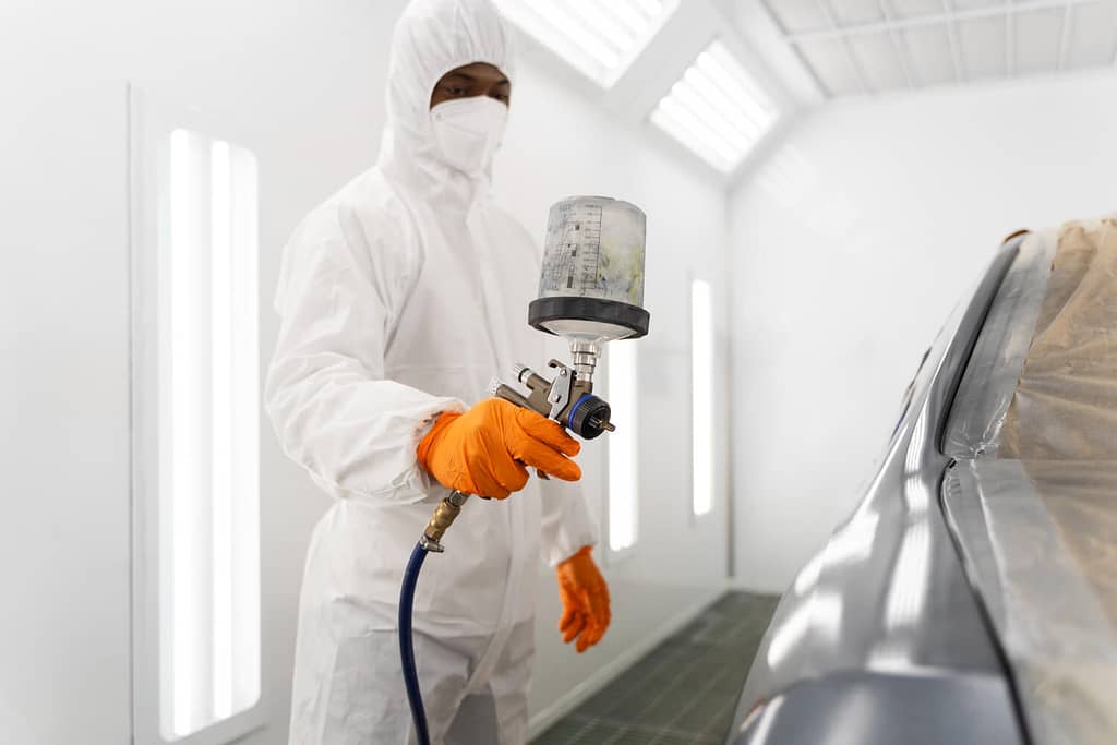 Aplicação de tinta em ambiente controlado com cabine pressurizada. Operador devidamente paramentado com EPI completo (macacão, máscara, luvas). Iluminação uniforme e paredes limpas indicam conformidade com boas práticas de pintura automotiva. - Laudo Cabine Pintura.