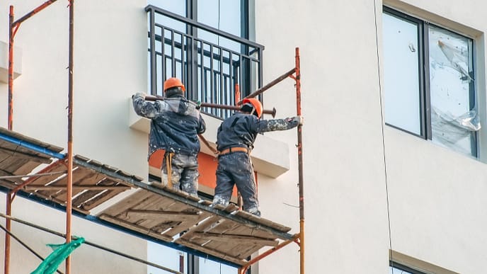Trabalhadores realizam montagem de andaime em fachada de edifício, atuando em altura com uso de capacetes e vestimentas de proteção - Curso Acesso por Corda 2 NBR 15475