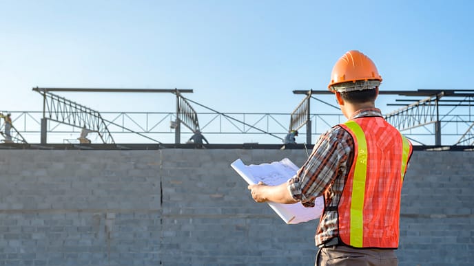 Um engenheiro ou supervisor de obra analisa plantas arquitetônicas em frente a um edifício em construção, assegurando que todos os detalhes estejam alinhados com o planejamento. - Curso Capacitação Incial NR 18. Curso NR 18 Inicial.